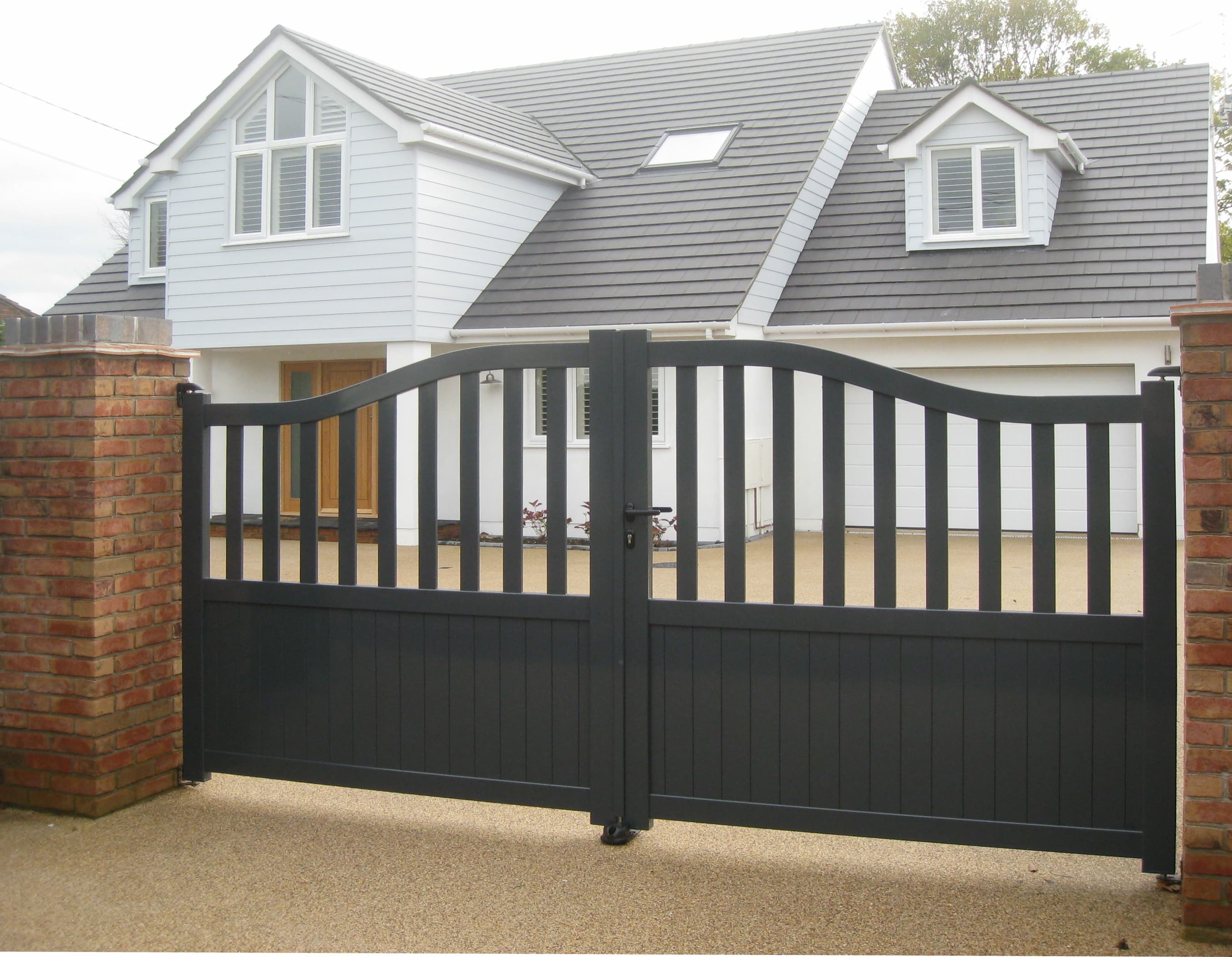 Charcoal grey aluminium driveway gate with curved top design, juxtaposed with brick pillars, in front of a modern white house with slate roofing and dormer windows.